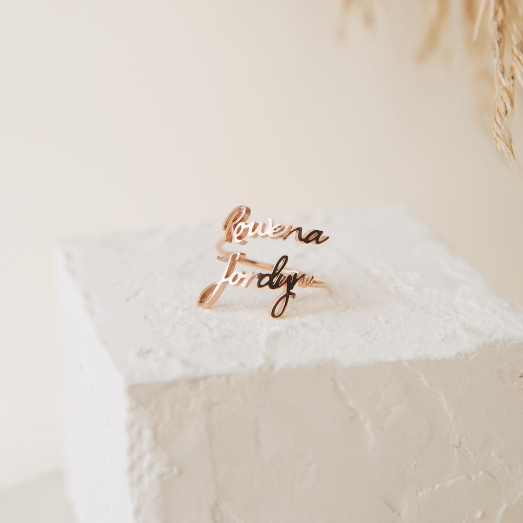 Two Grace Duo Name Rings in gold, featuring elegant cursive lettering, are displayed on a textured white block with a soft background, highlighting the charm of this personalized design.