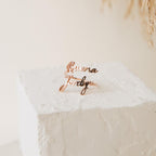 Two Grace Duo Name Rings in gold, featuring elegant cursive lettering, are displayed on a textured white block with a soft background, highlighting the charm of this personalized design.