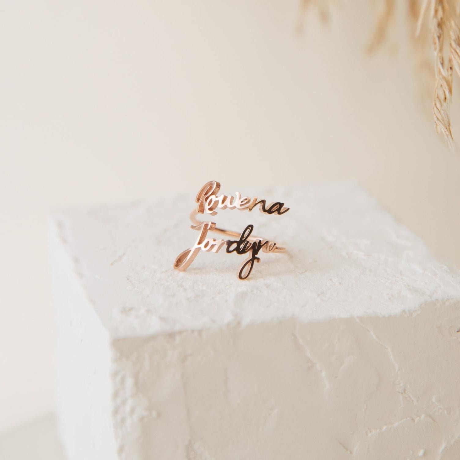 Two Grace Duo Name Rings in gold, featuring elegant cursive lettering, are displayed on a textured white block with a soft background, highlighting the charm of this personalized design.