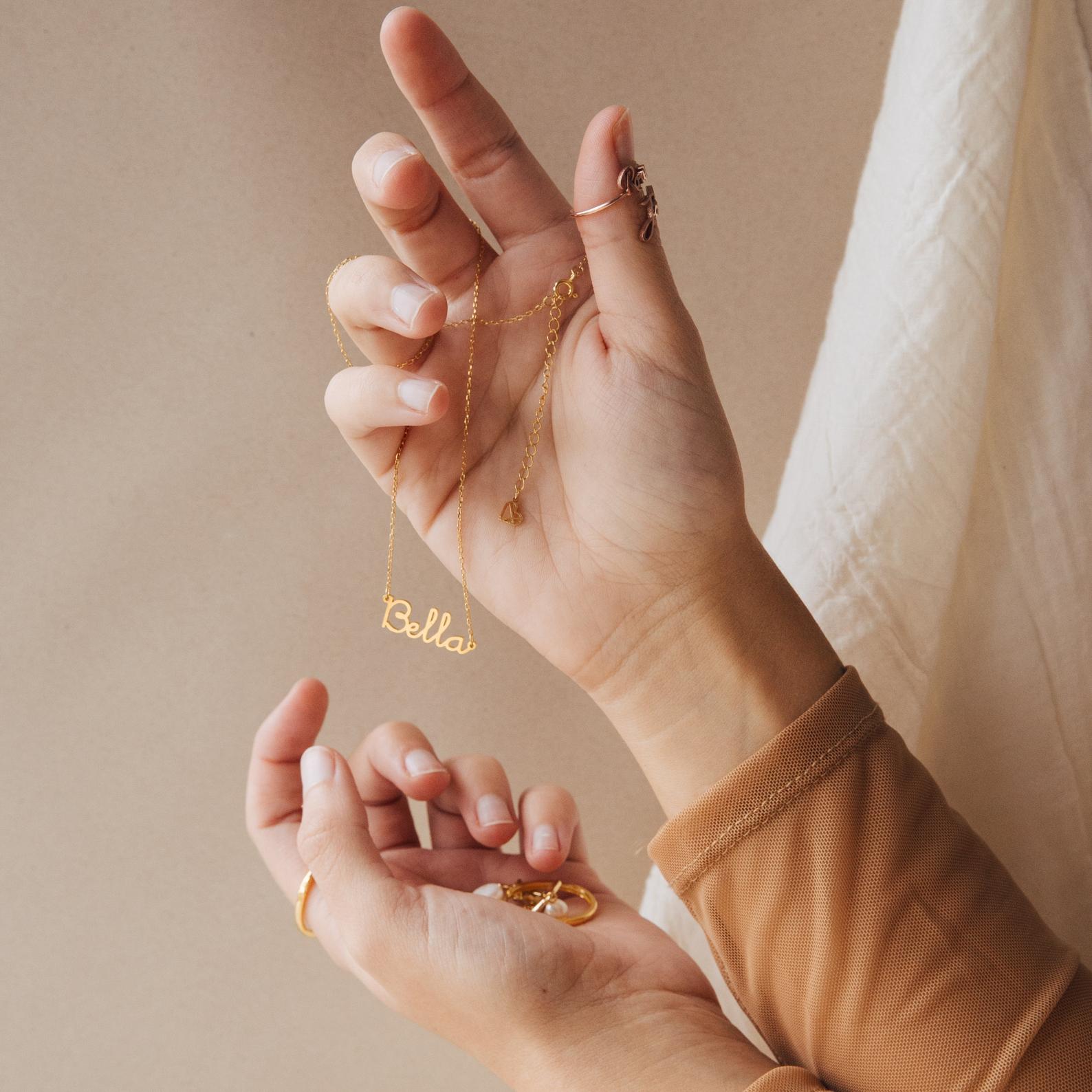 A hand holds the Dainty Name Necklace with "Bella" against a beige background and white cloth. This personalized necklace is the ideal minimalist jewelry piece for any occasion.