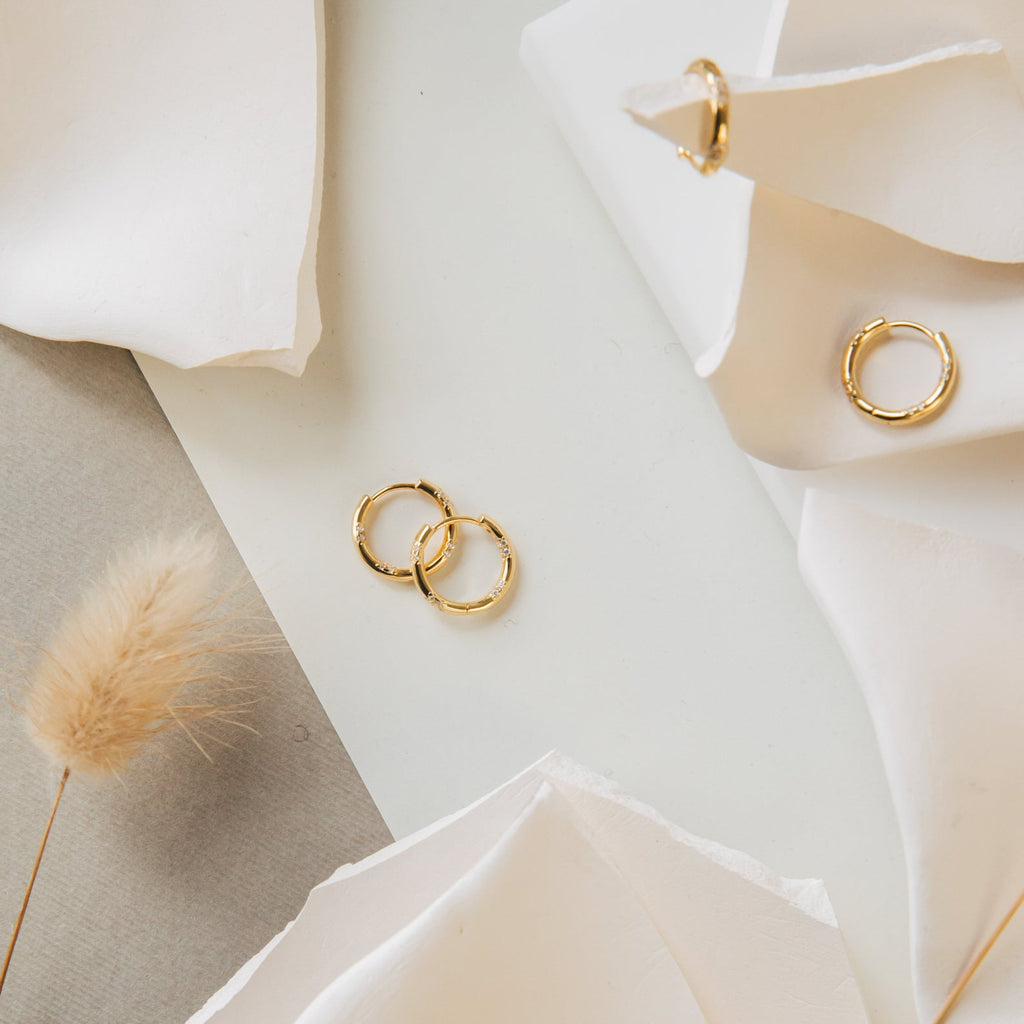 Kaia Diamond Huggies and gold hoop earrings displayed on a white surface with soft paper and dried plants, combining for a look of effortless elegance.