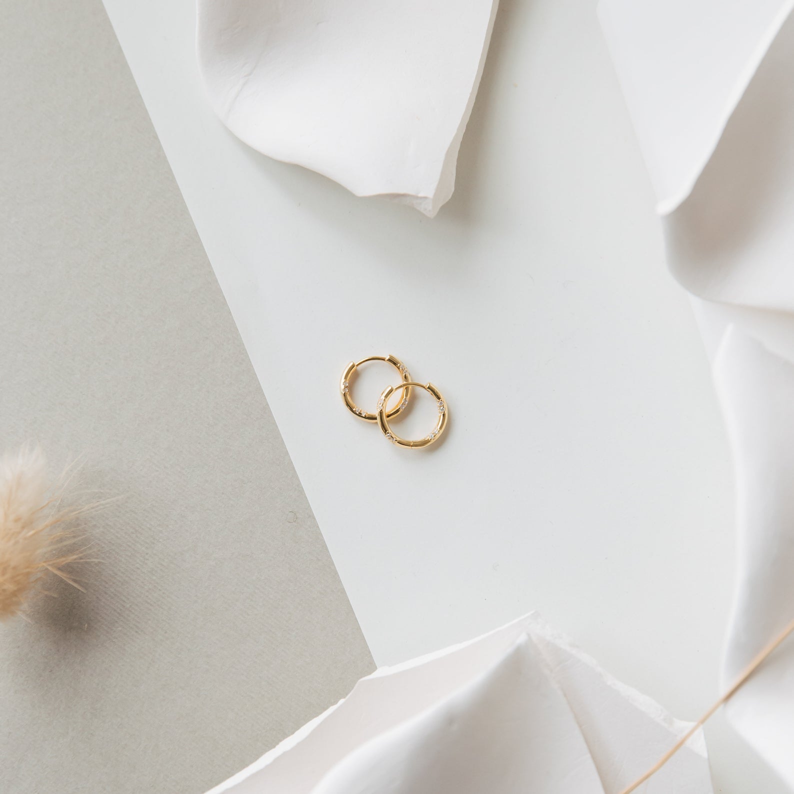 Two golden hoop earrings rest on a white and beige textured surface, surrounded by soft, neutral decorations—a timeless look that pairs beautifully with dainty diamond earrings or classic Kaia Diamond Huggies.