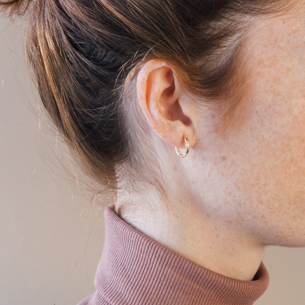 Close-up of a woman's ear adorned with Kaia Diamond Huggies, her hair tied up and a mauve turtleneck highlighting the earrings' elegant sparkle.