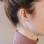 Close-up of a woman's ear adorned with Kaia Diamond Huggies, her hair tied up and a mauve turtleneck highlighting the earrings' elegant sparkle.