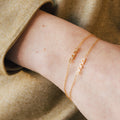 Two Petite Tiny Name Bracelets adorn a wrist, each gold chain featuring a delicate pendant: one inscribed with "mama," the other with "anna.