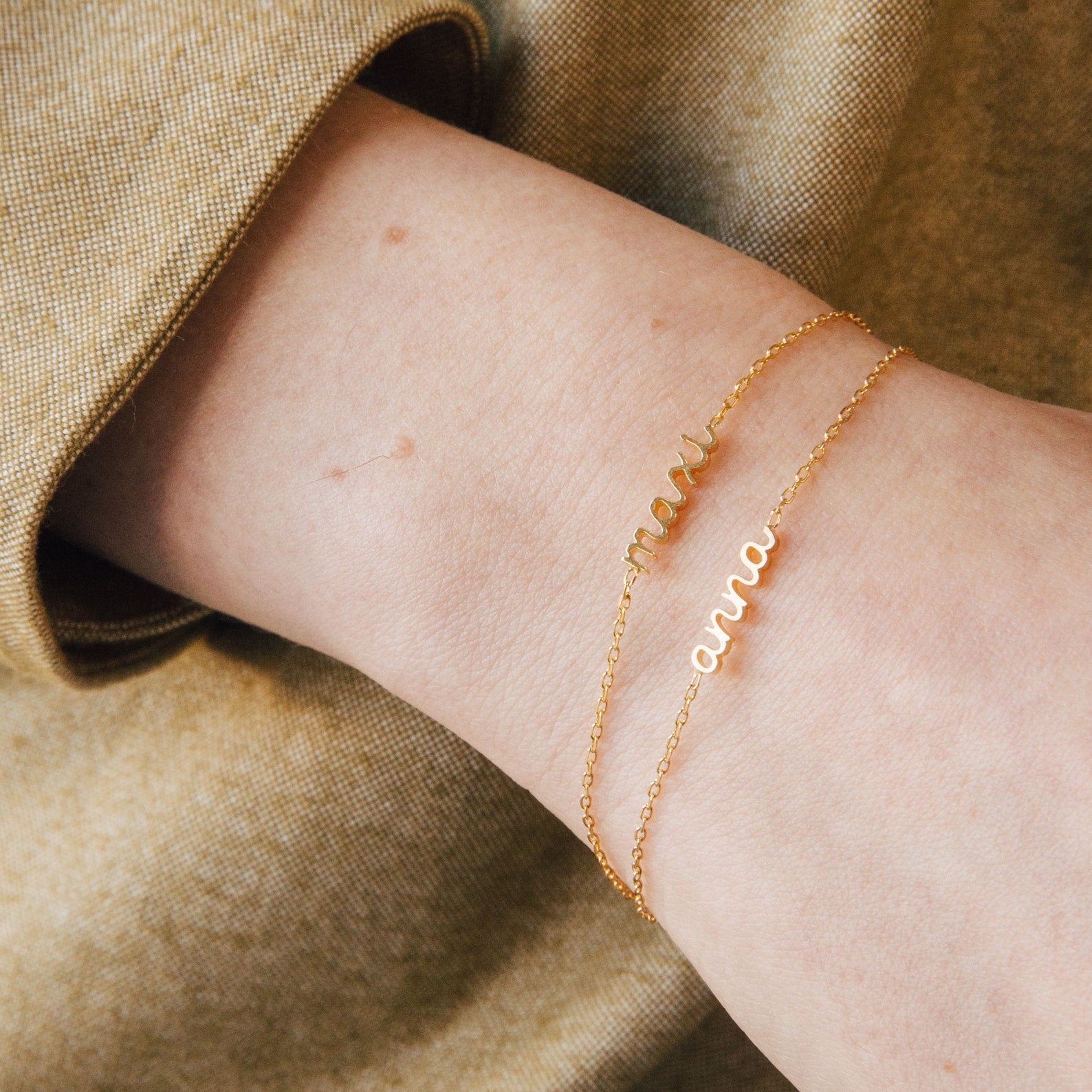 Two Petite Tiny Name Bracelets adorn a wrist, each gold chain featuring a delicate pendant: one inscribed with "mama," the other with "anna.
