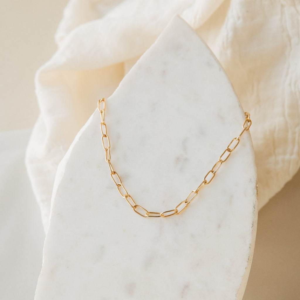 The Paperclip Toggle Necklace, featuring elongated gold links, is displayed on a white marble slab and soft beige fabric—ideal for boho-chic layered necklace styling.