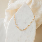 The Paperclip Toggle Necklace, featuring elongated gold links, is displayed on a white marble slab and soft beige fabric—ideal for boho-chic layered necklace styling.