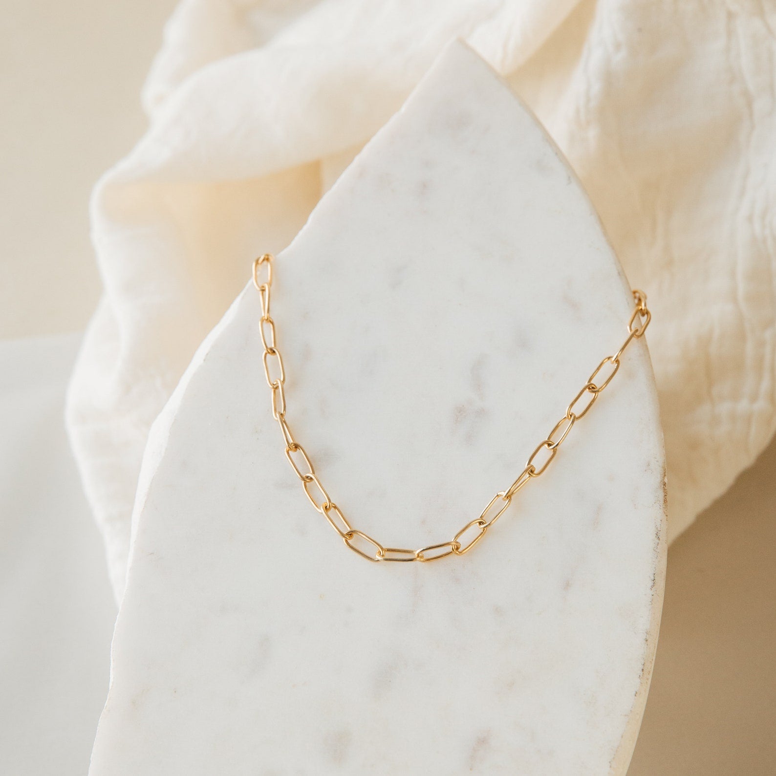 The Paperclip Toggle Necklace, featuring elongated gold links, is displayed on a white marble slab and soft beige fabric—ideal for boho-chic layered necklace styling.