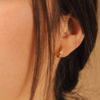 A close-up of a woman's ear wearing a dainty gold hoop earring featuring a single square-shaped-cut diamond at the center. 
