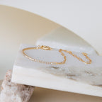The Klara Tennis Bracelet features a thin gold chain with sparkling stones, shown on a white stone slab against a neutral background for a minimalist, elegant look.