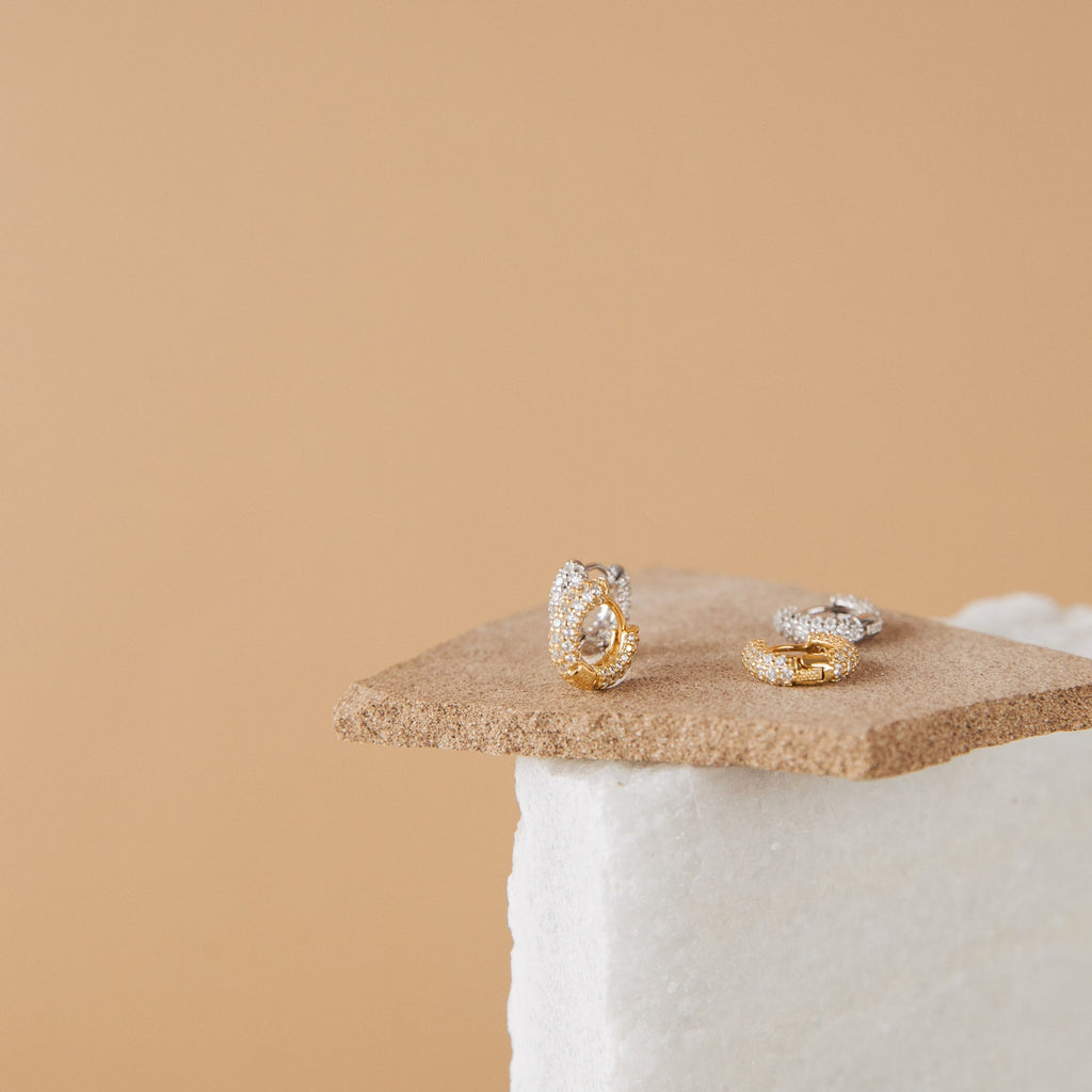 Kira Pave Huggies in gold and silver, adorned with small diamonds, are shown on textured beige and white backgrounds for an elegant look.