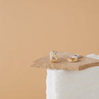 Kira Pave Huggies in gold and silver, adorned with small diamonds, are shown on textured beige and white backgrounds for an elegant look.