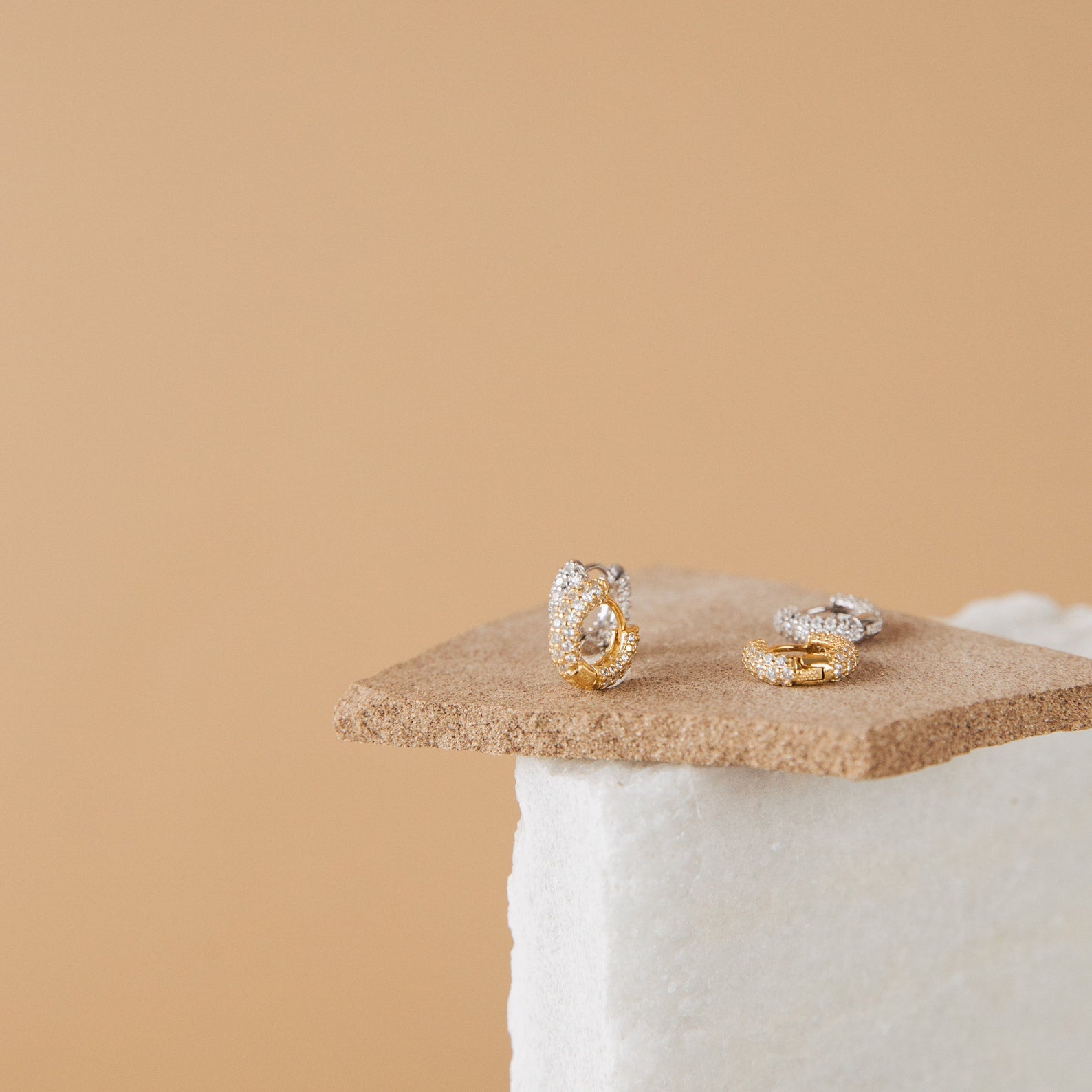 Kira Pave Huggies in gold and silver, adorned with small diamonds, are shown on textured beige and white backgrounds for an elegant look.