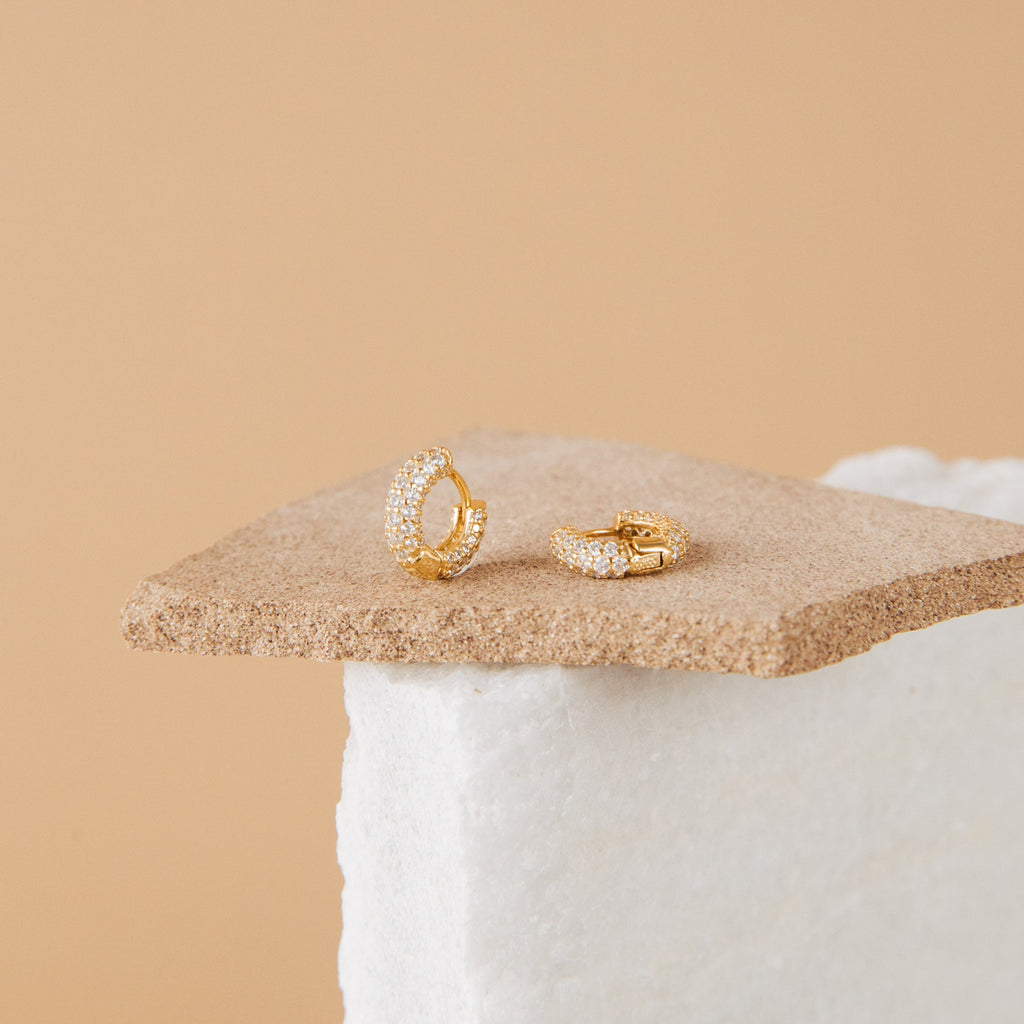 A pair of Kira Pave Huggies in small gold with pavé stones, displayed on a textured beige surface.