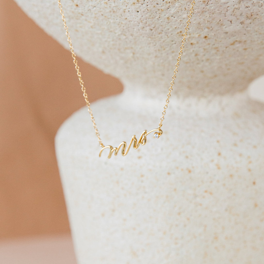 The Mrs. Necklace features a gold pendant spelling mrs in cursive, displayed on a textured white surface—an elegant wedding gift idea for the newlywed bride.