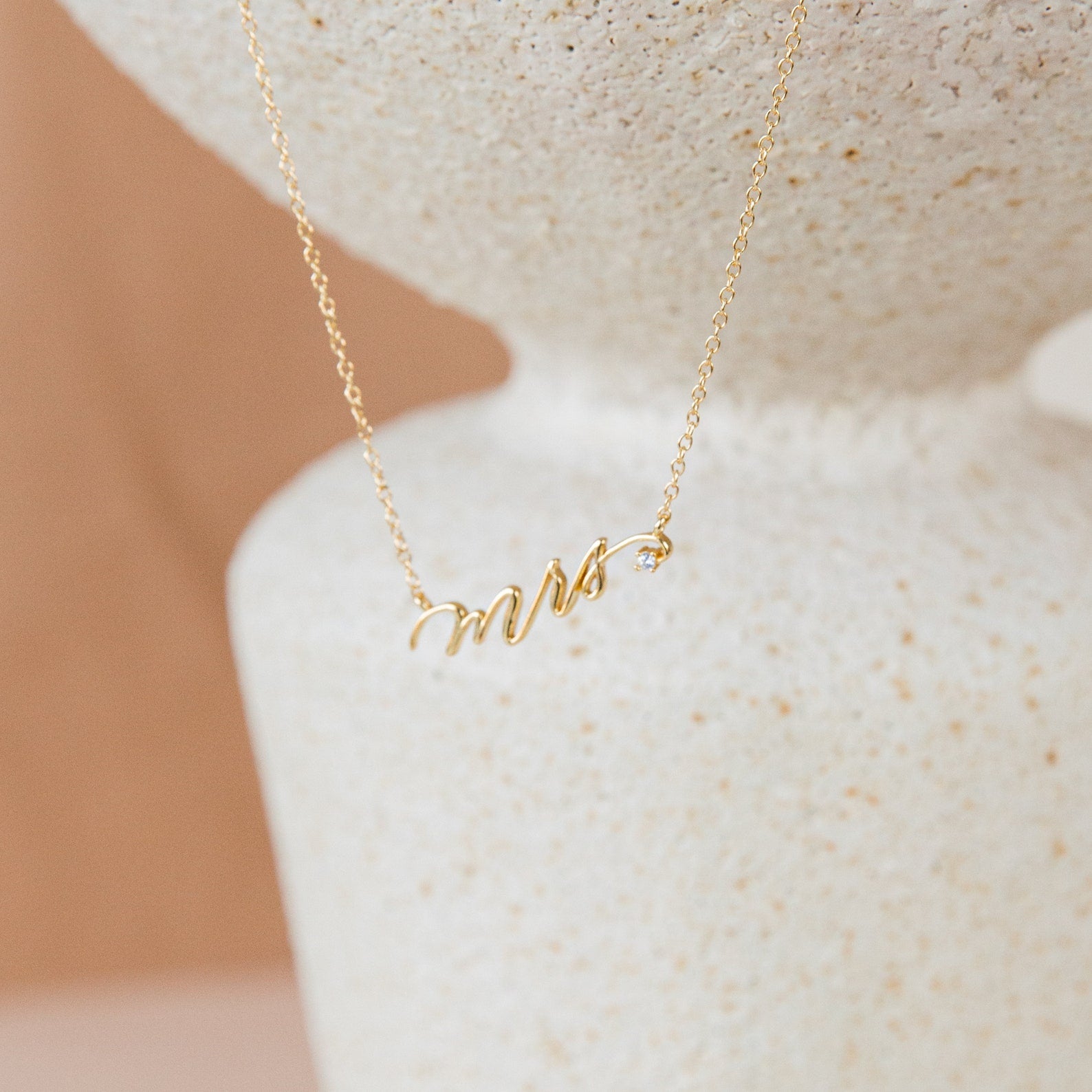 The Mrs. Necklace features a gold pendant spelling mrs in cursive, displayed on a textured white surface—an elegant wedding gift idea for the newlywed bride.