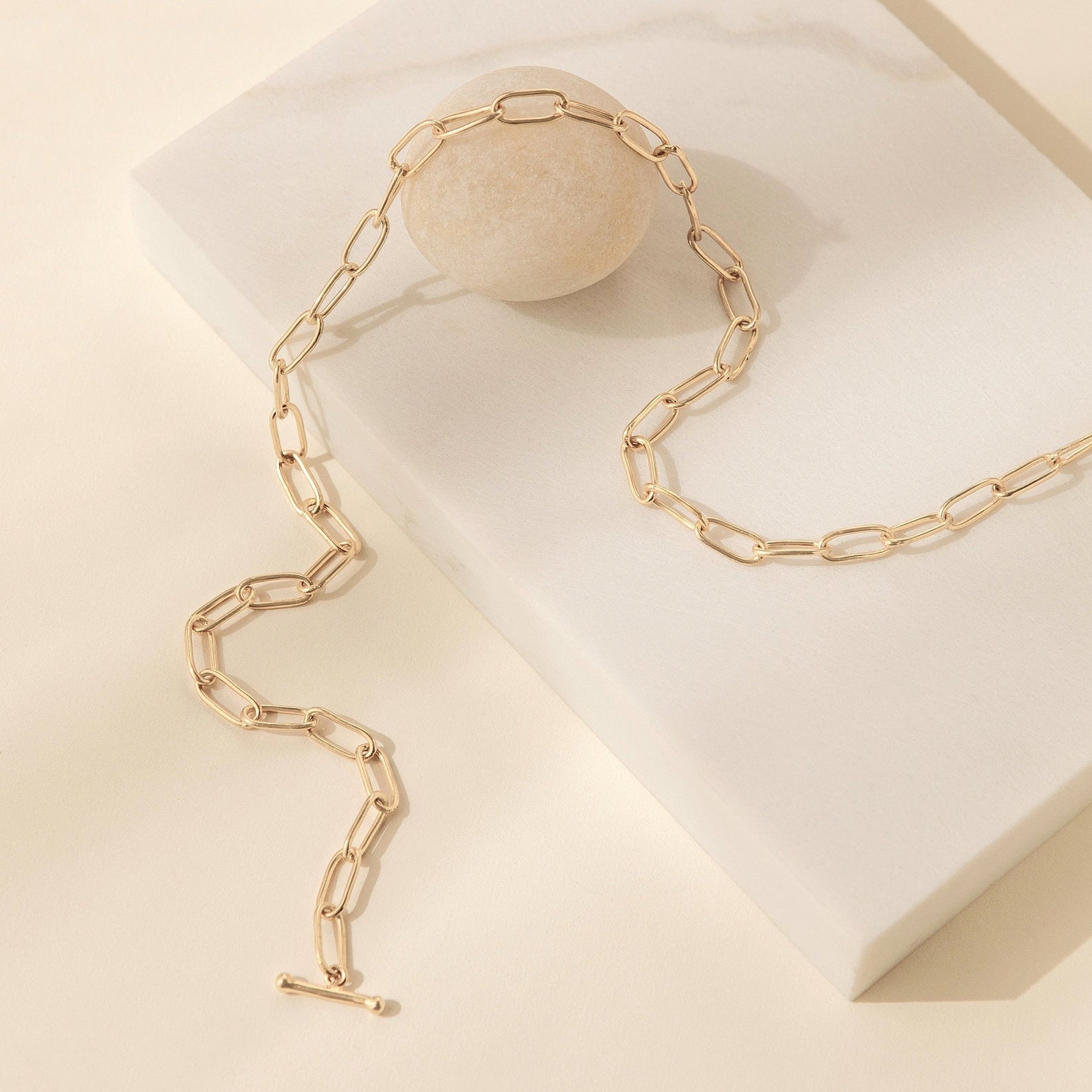 The Paperclip Toggle Necklace features a boho-chic gold chain with large links, showcased on a cream stone and beige background.