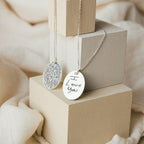 The Oval Fingerprint Necklace features two silver oval pendants—one with a fingerprint and the other engraved with "I love you"—resting on beige blocks, making it a meaningful piece of memorial jewelry ideal for personalized etching.