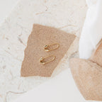 Two gold Ava Earrings displayed on a small brown textured paper—perfect statement pieces to add a bold touch to any look.
