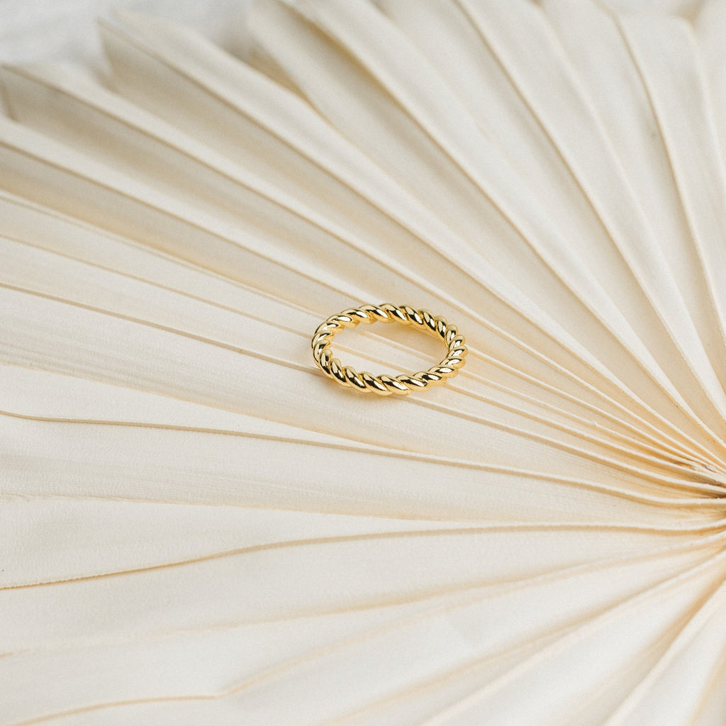 The Tia Twisted Ring in gold sits on a textured, cream fan-shaped surface—an elegant, modern piece ideal for stacking with your favorite rings.