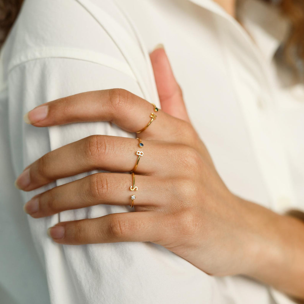 A hand displays three Modern Initial Birthstone Rings—customizable gold bands featuring small letters—worn against a white shirt.