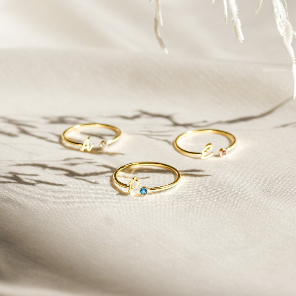Three gold Venice Initial Birthstone Rings rest on light fabric, each with a delicate gemstone and cursive lowercase letter charm—a personalized ring, perfect as an initial birthstone accessory for everyday elegance.