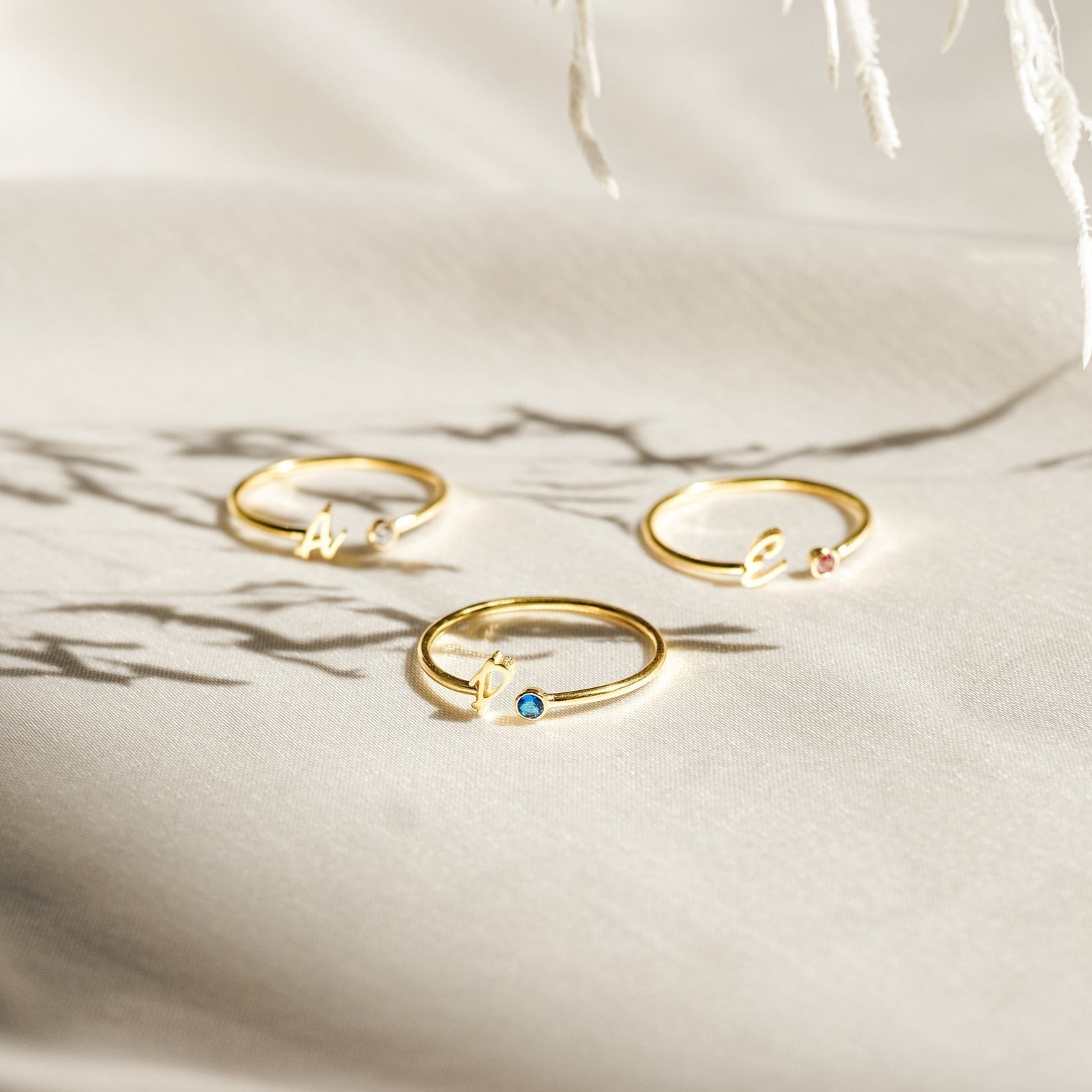 Three gold Venice Initial Birthstone Rings rest on light fabric, each with a delicate gemstone and cursive lowercase letter charm—a personalized ring, perfect as an initial birthstone accessory for everyday elegance.