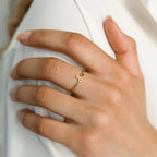 A manicured hand wears the Venice Initial Birthstone Ring—a delicate gold swirl band adorned with a small green gemstone and personalized initial charm.