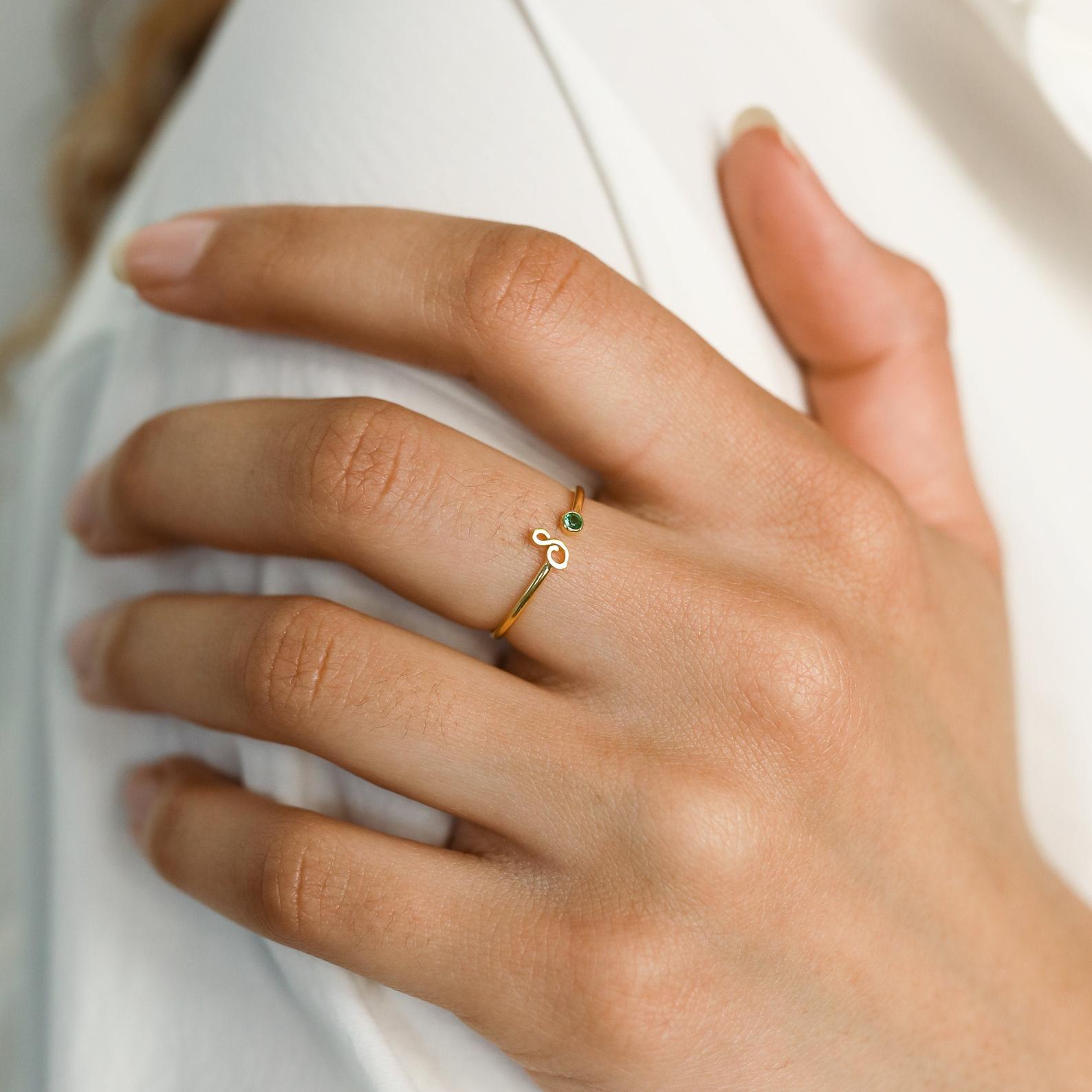 A manicured hand wears the Venice Initial Birthstone Ring—a delicate gold swirl band adorned with a small green gemstone and personalized initial charm.