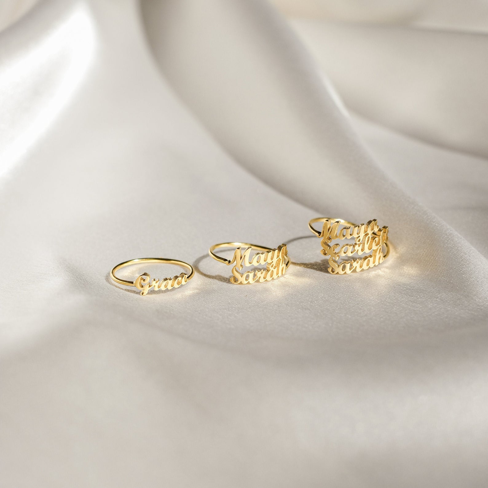 A gold London Duo Name Ring is elegantly displayed on soft cream-colored satin fabric—an exquisite example of personalized custom jewelry.