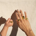 An adult and a child’s hand, each wearing gold rings—one the Initial Signet Ring—rest on light textured fabric in sunlight.