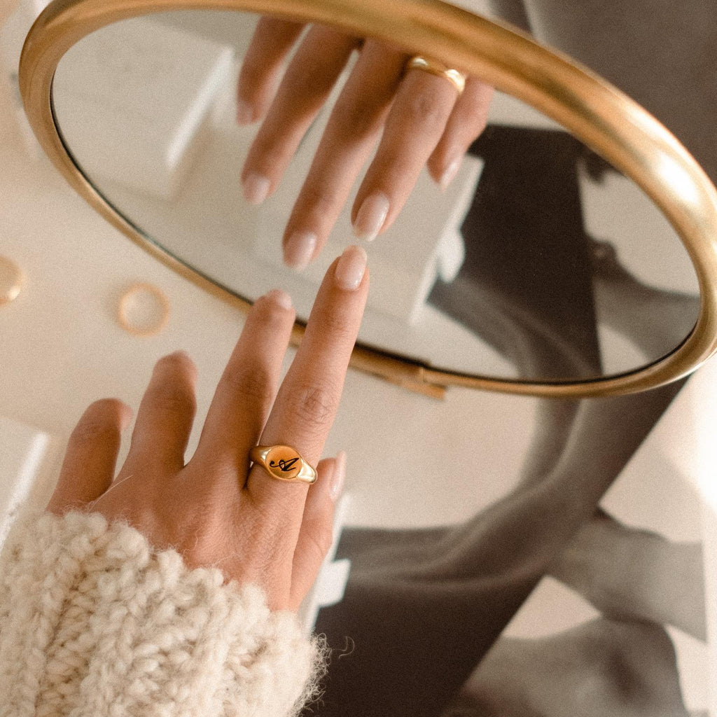 A hand wearing the Initial Signet Ring touches its reflection in a round mirror against a monochrome photo backdrop.