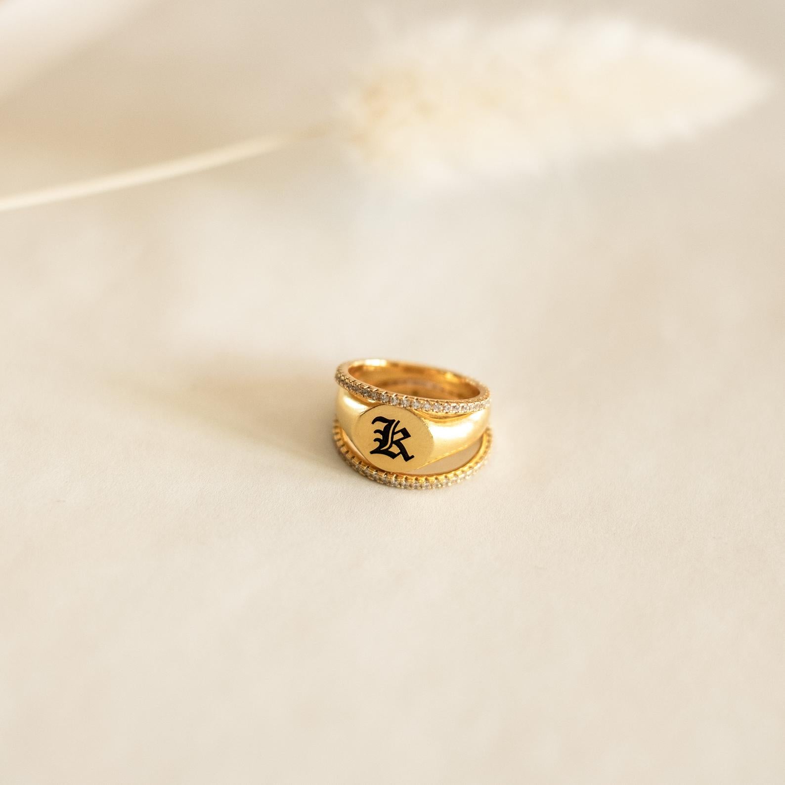 The Initial Signet Ring features a gold band with a black gothic-style K engraving, showcased on a softly blurred neutral background.