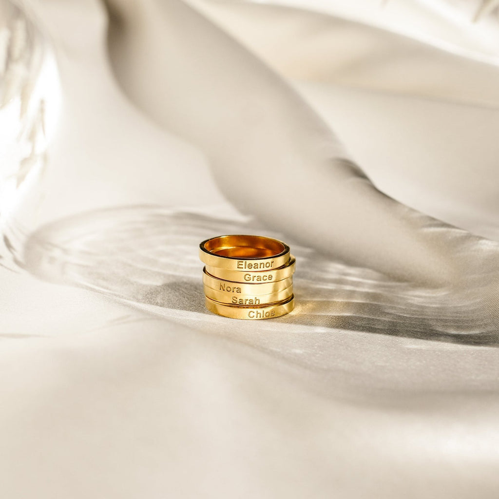 Four Alice Thin Engraved Rings, each personalized with a name, are stacked on soft ivory fabric in elegant lighting—a beautiful minimal ring set and the perfect custom gift.