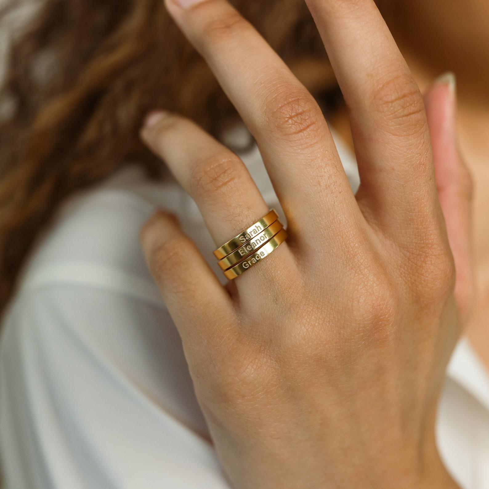 A hand wearing three Alice Thin Engraved Rings on the middle finger, each customized with a unique name—a meaningful personalized gift.