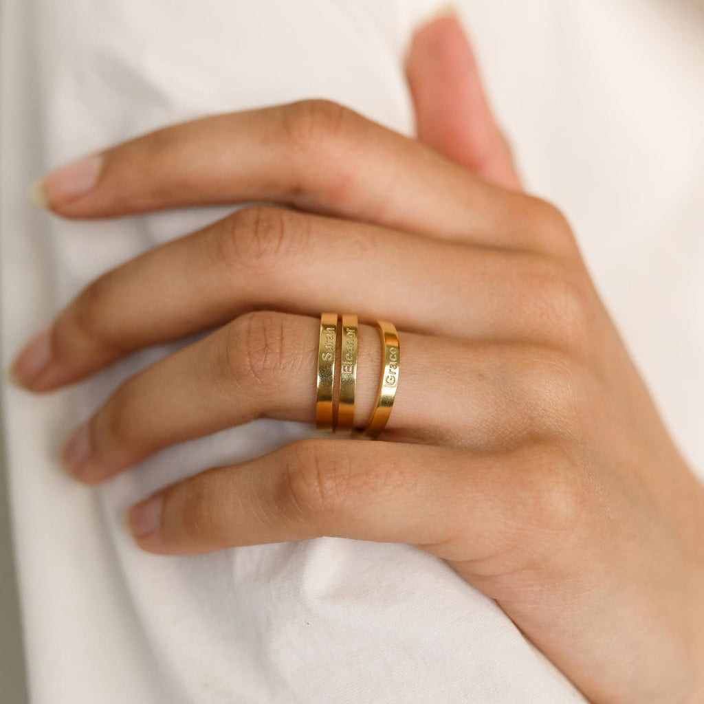 A hand adorned with four minimal rings, including the Alice Thin Engraved Ring, rests gently on white fabric.
