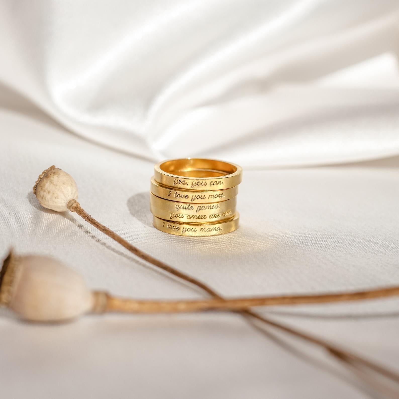 The Ann Thin Message Rings in rose gold, engraved with custom words, are stacked on white fabric beside dried poppy pods—a unique personalized ring set ideal for holiday gifting.