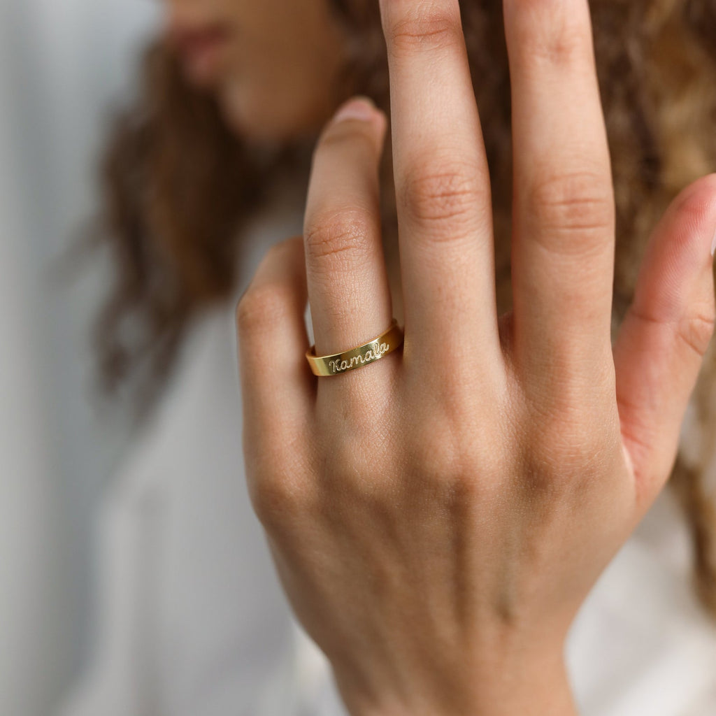 A person wearing the Ann Engraved Name Ring on their finger, with their face blurred in the background, showcases this timeless piece of jewelry.