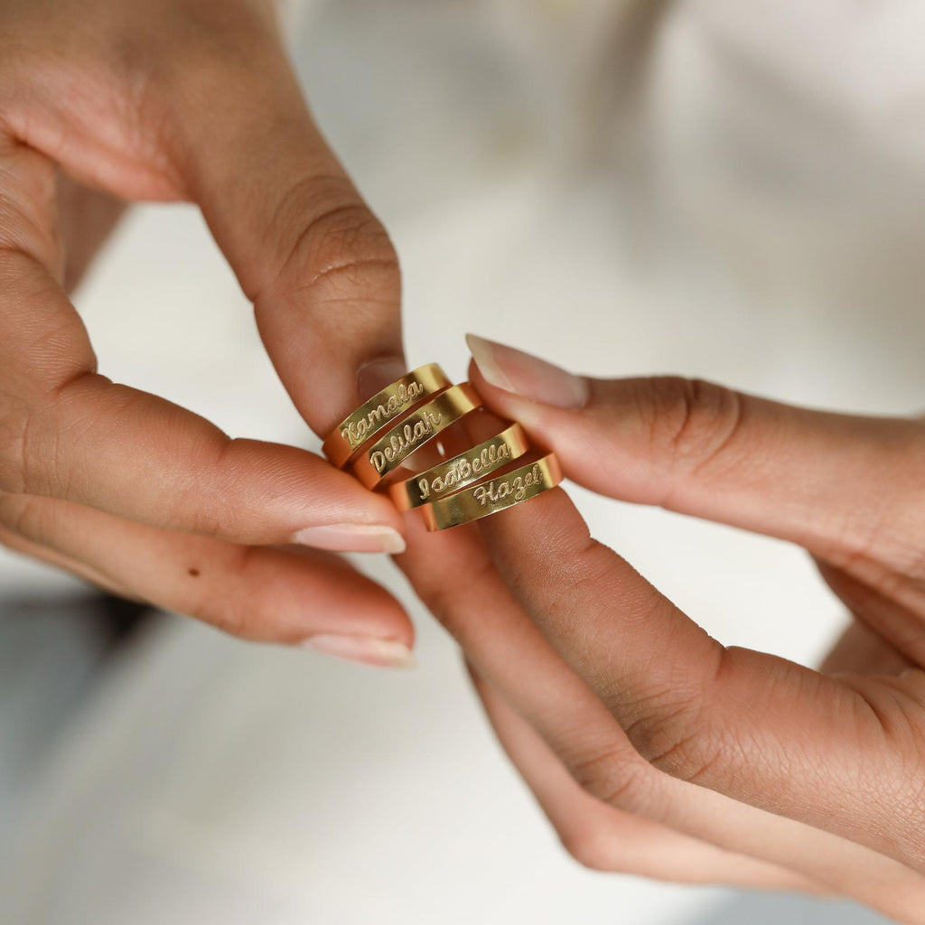 Close-up of hands holding two Ann Engraved Name Rings with delicate inscriptions, set against a soft neutral background—showcasing the timeless beauty of personalized jewelry.