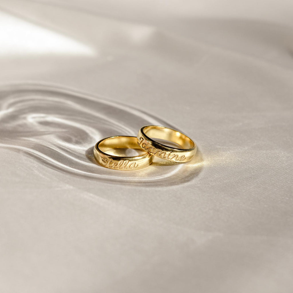 Two Ann Half Round Rings engraved with the names Stella and Loraine rest on soft, light fabric. Each personalized band highlights timeless elegance and meaning.