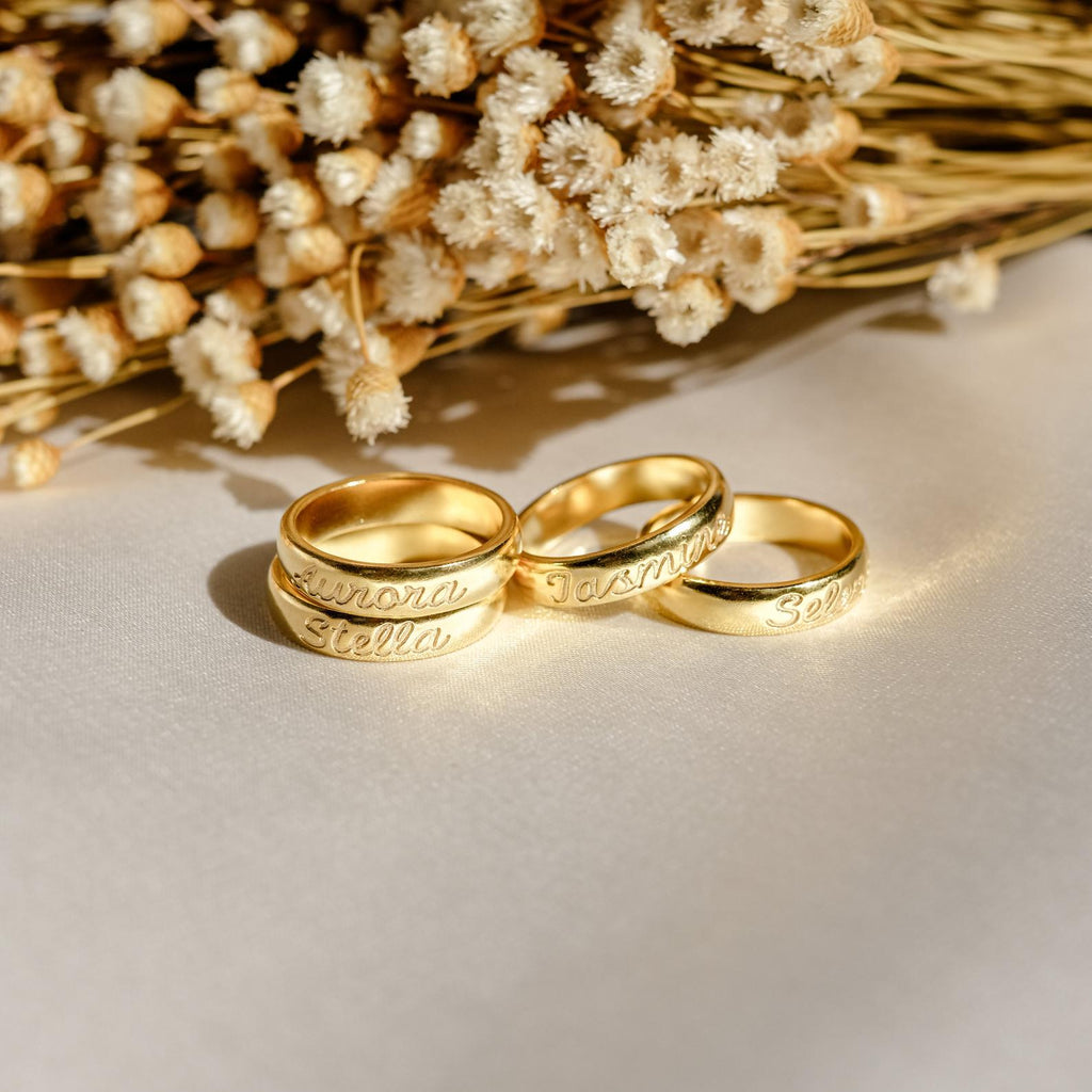 Four Ann Half Round Rings with personalized engravings rest on a light surface beside dried white flowers, each classic gold band capturing unique memories in elegant detail.