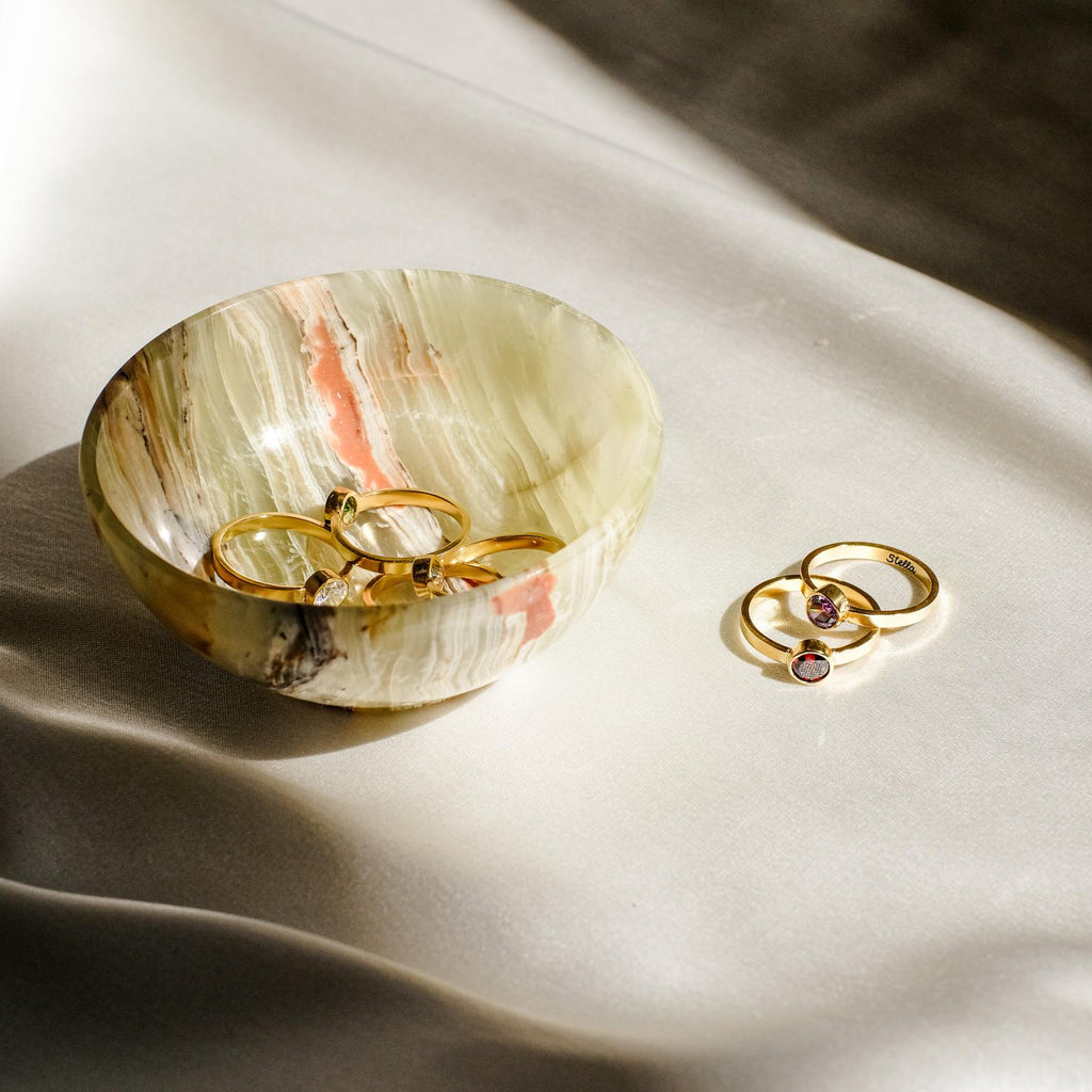 A marble bowl holds gold rings; the Birthstone Signet Ring rests nearby on silky fabric.