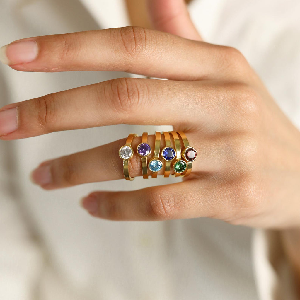 A hand wearing six gold rings, including the Birthstone Signet Ring and a vintage piece, each featuring a unique colored gemstone, shown against a neutral background.