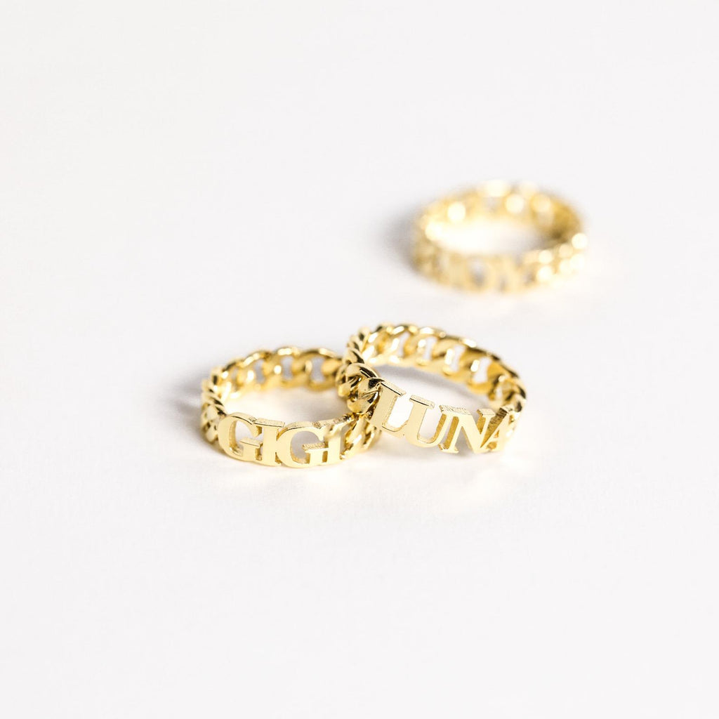 Three Newsletter Curb Name Rings with engraved letters rest on a white surface, two in front and one behind, highlighting their classic chain design—ideal for a personalized touch.