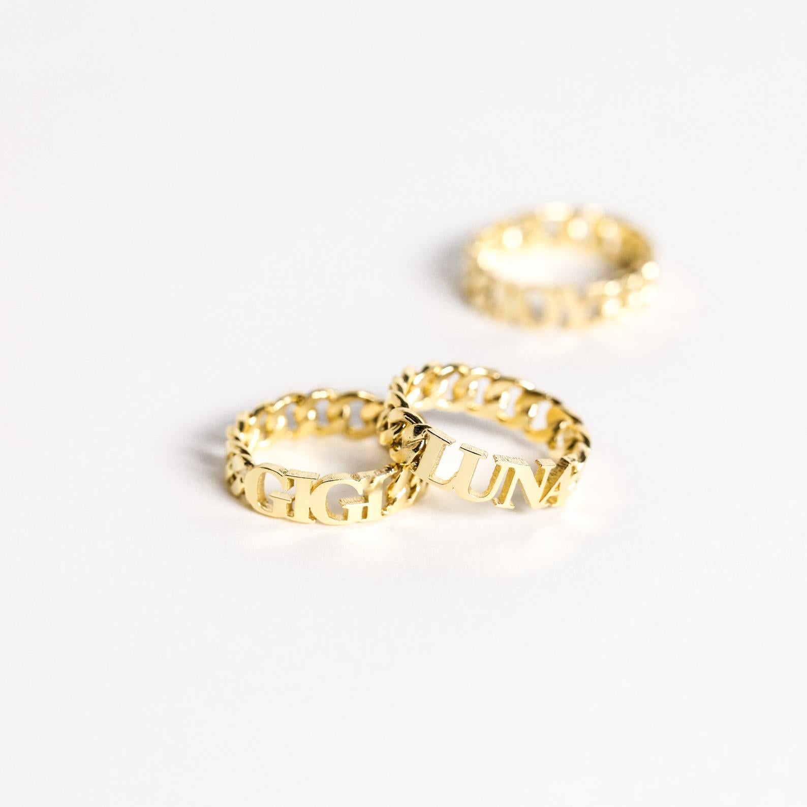 Three Newsletter Curb Name Rings with engraved letters rest on a white surface, two in front and one behind, highlighting their classic chain design—ideal for a personalized touch.
