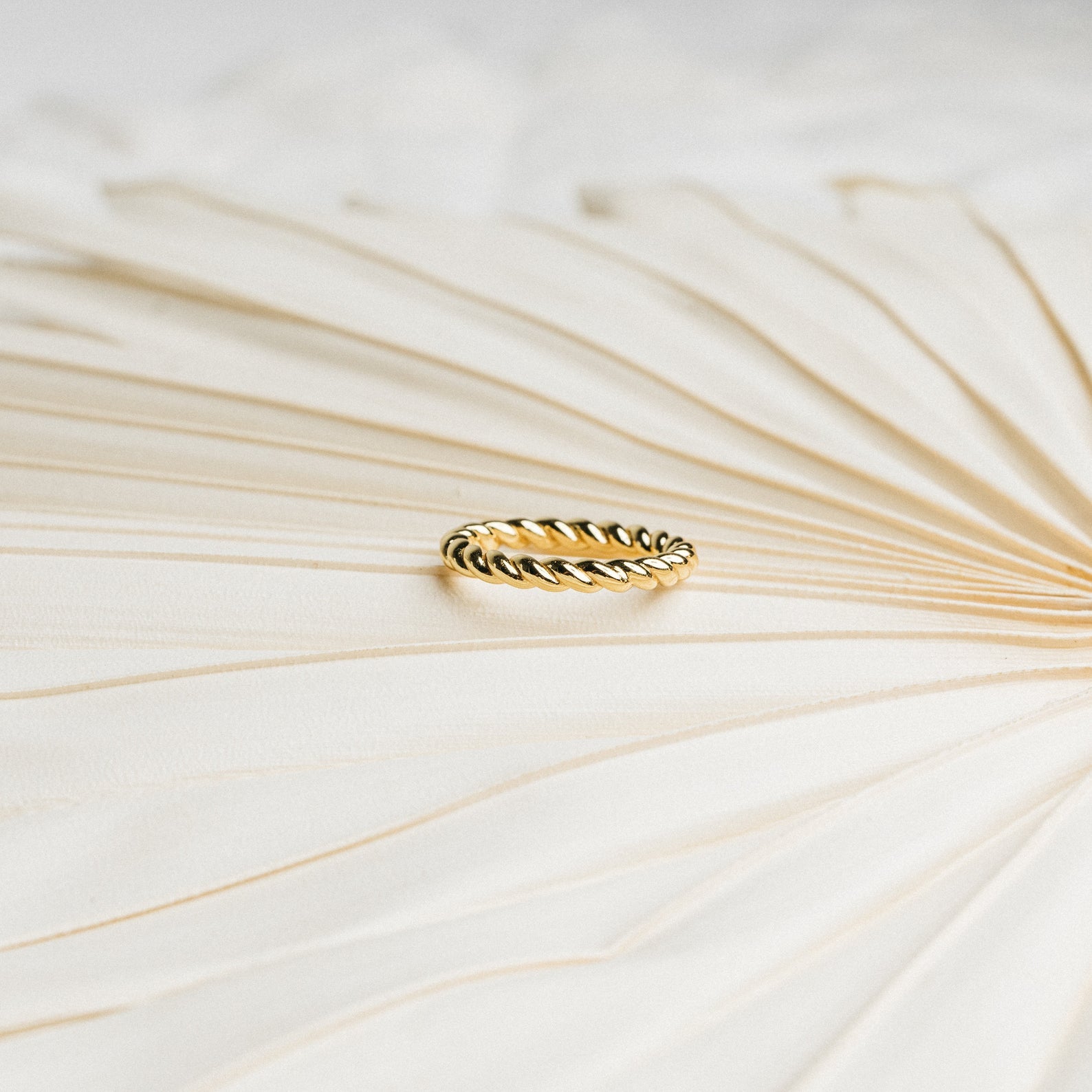 The Tia Twisted Ring in gold is shown on a textured, cream-colored surface with soft lighting—perfect for adding a modern twist to your ring stack.