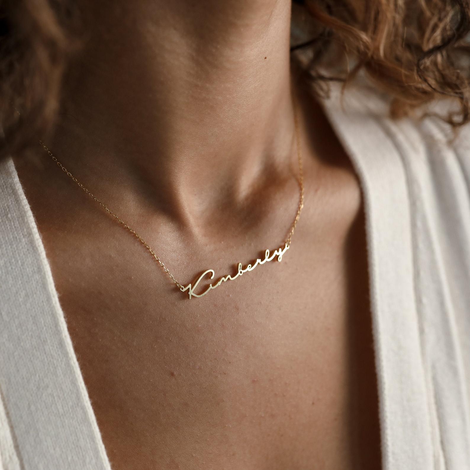 A woman wears a gold Minimal Name Necklace with a cursive "Kimberly" pendant and a cream top—the ideal personalized gift.