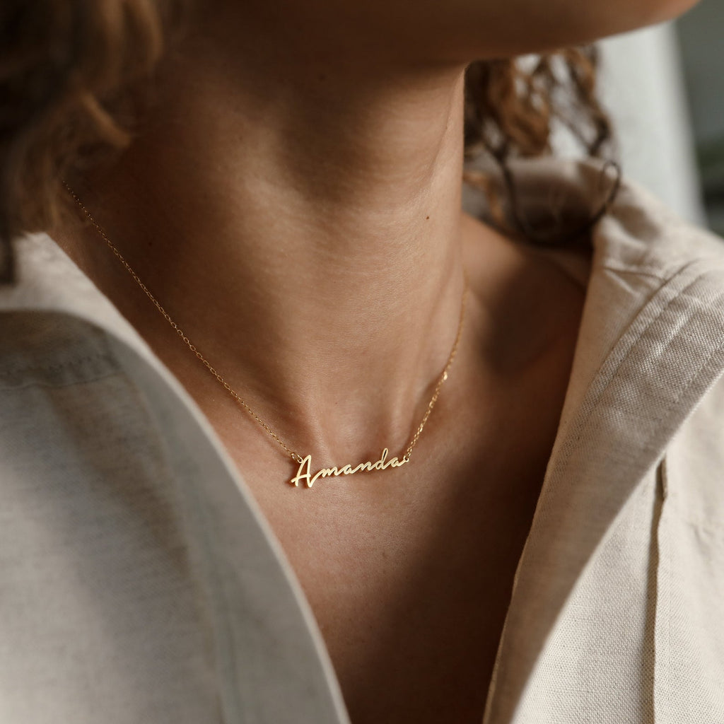 The Venice Name Necklace, featuring the name "Amanda" in a gold script font, is styled with a beige shirt for an elegant, dainty jewelry look.