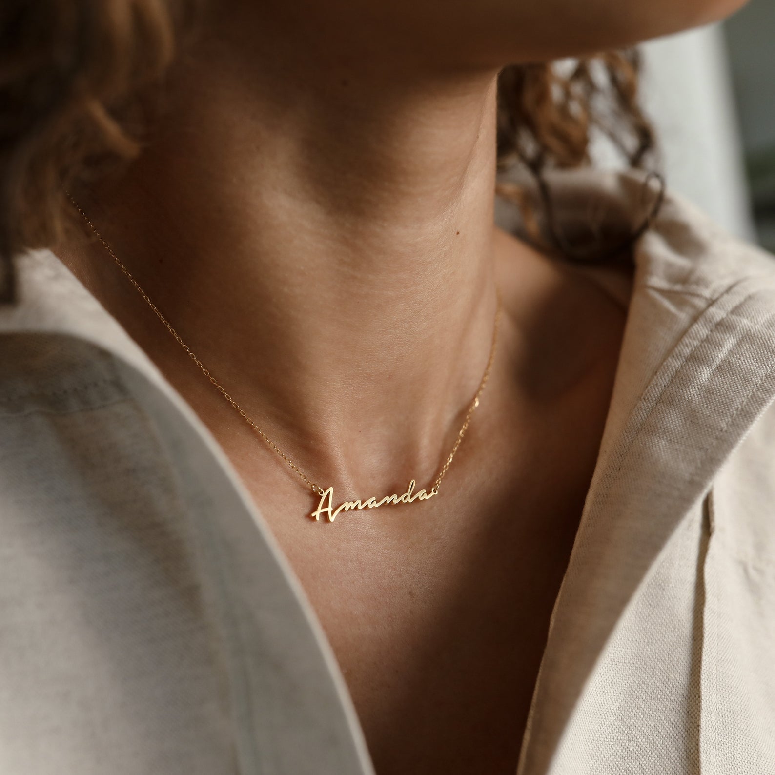 The Venice Name Necklace, featuring the name "Amanda" in a gold script font, is styled with a beige shirt for an elegant, dainty jewelry look.
